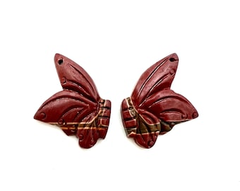 May include: Two carved butterfly wing ornaments. The wings are a deep reddish-brown color with detailed veining and a textured surface. The base of the wings has a striped pattern of brown, green, and gold.