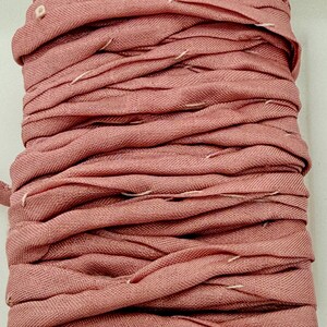 May include: A spool of dusty rose-colored ribbon. The ribbon is made of a woven fabric and is tightly wound. White stitching is visible along the edges of the ribbon, adding a subtle contrast to the overall color. The ribbon is likely used for crafting or decorative purposes.