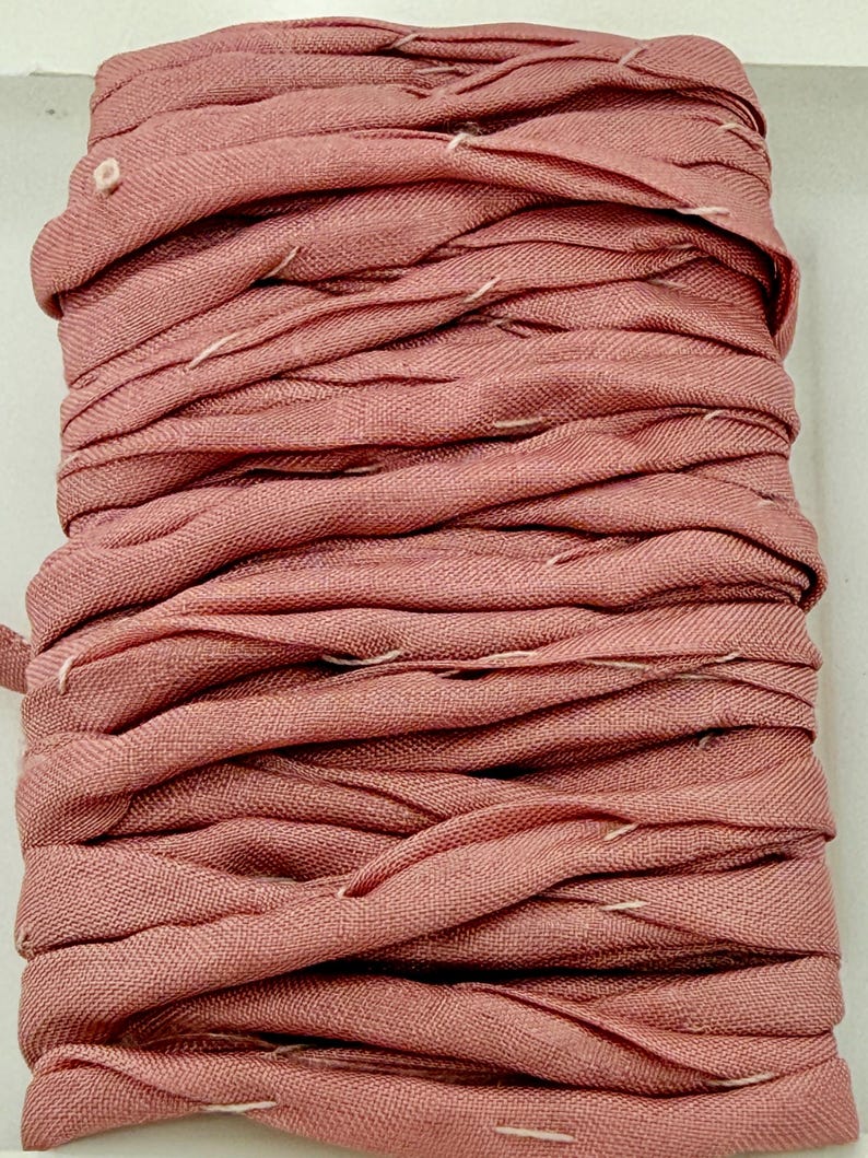 May include: A spool of dusty rose-colored ribbon. The ribbon is made of a woven fabric and is tightly wound. White stitching is visible along the edges of the ribbon, adding a subtle contrast to the overall color. The ribbon is likely used for crafting or decorative purposes.