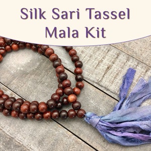 May include: A wooden mala kit with a silk sari tassel. The mala beads are a deep reddish-brown color. The tassel is a mix of blue and purple hues. The text "Silk Sari Tassel Mala Kit" is at the top of the image.