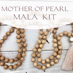 May include: Two wooden bead malas with a large pearl bead and tassel. The malas are made of light brown wooden beads strung on white string. The text "MOTHER OF PEARL MALA KIT" is at the top of the image.