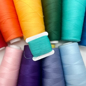 May include: A collection of colorful thread spools in a rainbow of hues, including red, orange, yellow, green, teal, blue, and purple. The spools are arranged closely together, showcasing the variety of colors available for sewing and crafting projects.