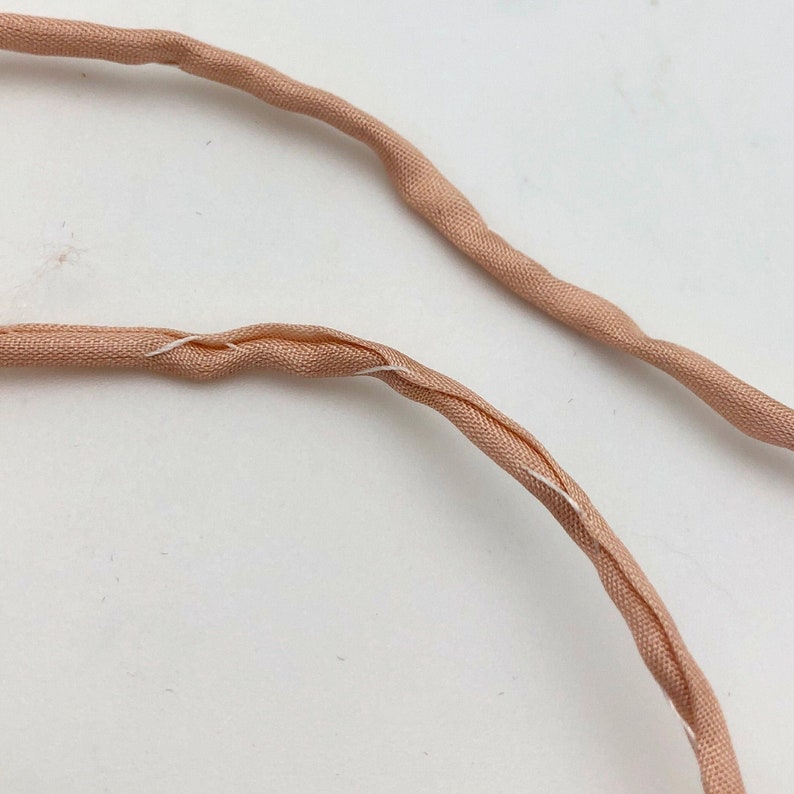 May include: Close-up of two strands of peach-colored fabric cord. The cords are twisted and have a slight sheen. White thread is visible where the fabric is joined. The background is a plain white surface.