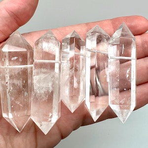 May include: Five clear quartz crystal points with pointed tips. The crystals are held in a hand and appear to be polished. The crystals are transparent with some internal inclusions. These crystals are often used for healing and meditation.