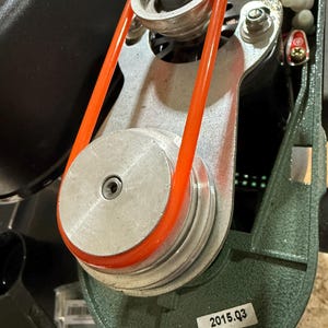 May include: Close-up of a machine component featuring an orange belt wrapped around multiple silver pulleys. The assembly is mounted on a green metal frame. A label with "2015.Q3" is visible.