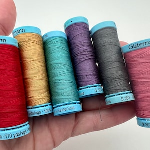 May include: A hand holding six spools of Gütermann thread in various colors: red, gold, teal, purple, gray, and pink. Each spool has a blue base and top with the brand name and thread details. The thread is 100% silk and 110 yards.