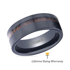 Puede incluir: Un anillo de titanio negro con una incrustación de madera marrón. El anillo está grabado con la palabra "TITANIUM".