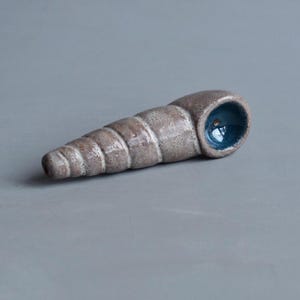 Könnte beinhalten: Eine Keramikpfeife in Form einer spiralförmigen Muschel mit einer texturierten, gesprenkelten hellbraunen Außenseite und einer glatten, glänzend tiefblauen Innenschale. Die längliche, konische Form weist deutliche Rillen und eine kleine Öffnung auf.