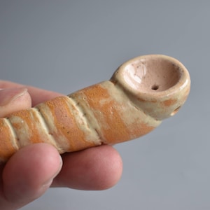 May include: A handmade ceramic pipe with a spiral design in shades of orange and cream. The bowl is a light pink color. The pipe is held in a person's hand.