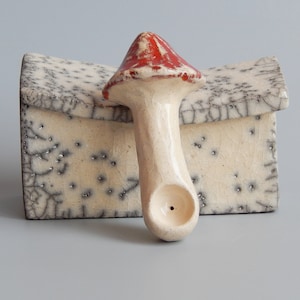 May include: A ceramic mushroom pipe with a red and white glaze. The pipe is sitting on a white ceramic box with a crackled glaze.