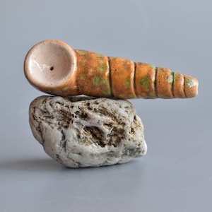 Puede incluir: Una pipa de cerámica en tonos naranja y verde descansa sobre una piedra gris. La pipa tiene forma cilíndrica con un cuenco en un extremo y un pequeño agujero. La piedra es rugosa y texturizada.