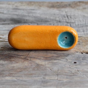 Puede incluir: Una pipa de cerámica lisa y alargada con un acabado exterior de color naranja vibrante y un interior de cazoleta circular en contraste esmaltado en azul turquesa brillante. El objeto presenta un pequeño orificio centrado en la base.