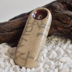 May include: A ceramic pipe with a crackle glaze finish. The pipe is beige with brown and black crackle lines. The bowl of the pipe is a dark red color.