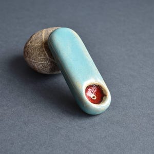 May include: A ceramic pipe with a light blue glaze and a red interior. The pipe is oval-shaped and rests on a small, textured stone. The background is a solid, dark gray.