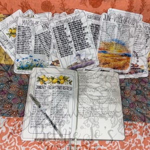 May include: An open journal with a pen, surrounded by several cards. The journal's cover reads "JANUARY-FRESH START REFRESH" with a lemon illustration. The cards display months and titles like "SEPTEMBER-LIGHT" and "JUNE-VOICE OF GOD". The background is a colorful patchwork quilt.