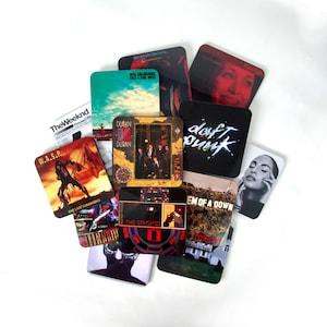 May include: A collection of square coasters featuring various music album cover art, including W.A.S.P., The Weeknd, Duran Duran, Daft Punk, The Strokes, and System of a Down. The coasters are arranged in a scattered pile, displaying diverse graphic designs.