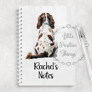May include: A white spiral-bound notebook featuring a watercolour illustration of a brown and white spaniel dog viewed from behind. The cover includes the text "Little Positive Things" inside a floral wreath and the personalised name "Rachel's Notes".