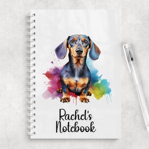 Puede incluir: Un cuaderno de espiral blanco con una colorida ilustración en acuarela de un perro salchicha negro y fuego. Debajo del perro, el texto Rachel's Notebook está impreso en una fuente cursiva negra. Un bolígrafo de plástico transparente está al lado.