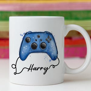 May include: White ceramic mug with a blue watercolour-style video game controller design. The name "Harry" is written in a black script font below the controller. The mug has a curved handle.