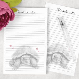 May include: Two white lined notepads with the text "Rachel's notes" in cursive. One notepad has a pencil drawing of a tortoise and small pink hearts. A clear pen rests on the second notepad. A pink flower is in the upper left corner.