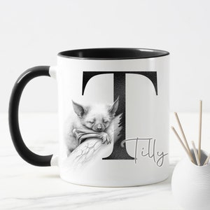 May include: A white ceramic mug with a black handle and rim. The mug features a black and white illustration of a bat sleeping on a branch, with the letter "T" behind it. The name "Tilly" is written below the bat.