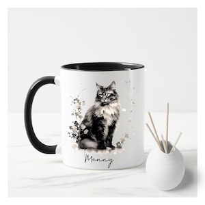 May include: White ceramic mug with a black interior and handle. The mug features a black and white illustration of a cat with the name "Manny" in script. A small white vase with wooden sticks is next to the mug.