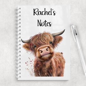 Pode incluir: Um caderno de espiral branco com o texto Rachel's Notes em letra cursiva preta e uma ilustração colorida de uma vaca Highland castanha e peluda em pé na relva alta. Uma caneta de plástico transparente está posicionada à direita do caderno.