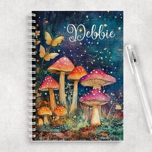 May include: A spiral-bound notebook featuring a dark, starry forest scene with colorful mushrooms and butterflies. The name Debbie is printed in white script at the top. A clear plastic pen with a silver clip rests beside the notebook.