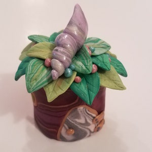 Puede incluir: Una pequeña caja decorativa con una tapa hecha de arcilla marmoleada verde y blanca. La tapa está decorada con una concha de mar púrpura y blanca, hojas verdes y cuentas rosadas y azules.