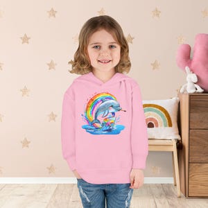 Puede incluir: Una sudadera con capucha rosa con un gráfico colorido de un delfín saltando a través de un arcoíris. El delfín sostiene un pincel y el diseño incluye salpicaduras de agua. La sudadera con capucha la lleva un niño pequeño.