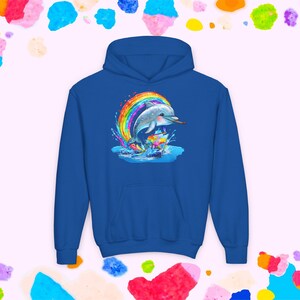 Puede incluir: Una sudadera con capucha azul real con un gráfico colorido de un delfín saltando a través del agua bajo un arcoíris. El delfín es gris y blanco, con un pequeño objeto marrón en la boca. El diseño está centrado en la parte delantera.