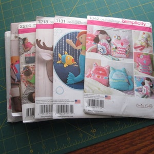 May include: Five different sewing patterns for making stuffed toys. The patterns are from Simplicity and include a mermaid, a reindeer, a fish, an owl, and a cat. The patterns are all on white paper with colorful illustrations.