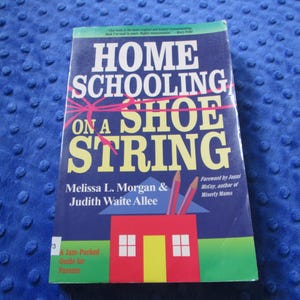May include: A book titled "Home Schooling on a Shoe String" with a blue cover. The cover features large white text, pink accents, and a graphic of a house and pencils. The book is a guide for parents.