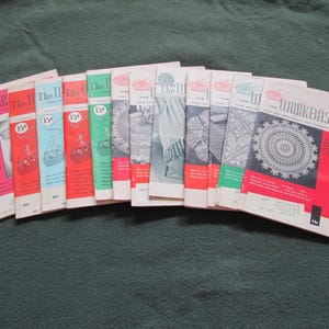 May include: A collection of vintage "Workbasket" magazines, featuring colorful covers with illustrations of crafts. The magazines are stacked on a green surface, showcasing a variety of designs and patterns.