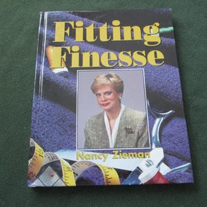 May include: A hardcover book titled "Fitting Finesse" with a woman's portrait on the cover. The cover is a mix of purple and blue hues, with yellow text and a measuring tape design. The author's name, Nancy Zieman, is also visible.