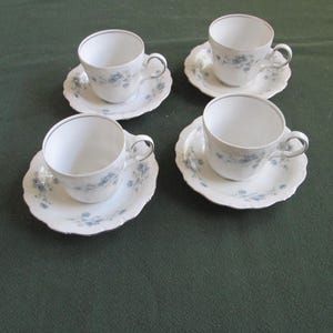May include: Four white teacups and saucers with a delicate blue floral pattern and silver trim. The cups have a classic handle design and sit on matching scalloped-edge saucers. A charming set for tea parties or display.