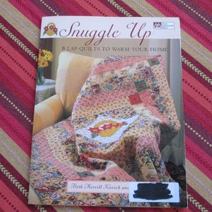 May include: A book cover titled "Snuggle Up" featuring a colorful quilt draped over a chair. The quilt has a floral pattern with pink, yellow, and green hues. The book promises "8 Lap Quilts to Warm Your Home."