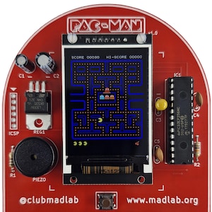 Könnte beinhalten: Eine rote Pac-Man-Spielkonsole mit einem Bildschirm, der das klassische Arcade-Spiel anzeigt. Die Konsole hat Knöpfe, einen Lautsprecher und verschiedene elektronische Komponenten. Der Text "PAC-MAN" steht oben, und die Website-Adresse ist sichtbar.