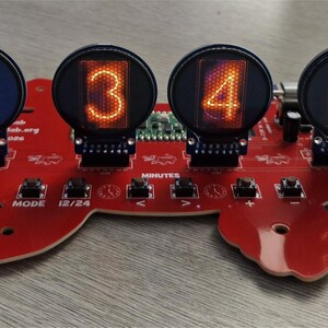 May include: A red turtle-shaped circuit board featuring six circular nixie tube displays showing the numbers one through six in glowing orange. The board includes several small black push buttons labeled with text such as MODE, 12/24, and symbols for time adjustment.