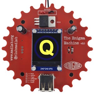 Op de afbeelding: Een rode printplaat met een klein LCD-scherm dat de letter "Q" in geel weergeeft. De printplaat is gelabeld met "The Enigma Machine v2.0" en heeft een USB-poort en verschillende knoppen. De printplaat is ook gelabeld met de website "www.madlab.org" en de social media-handle "@clubmadlab".