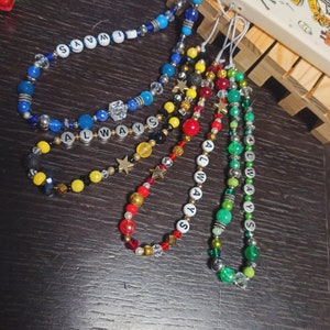 Może przedstawiać: Kolekcja koralikowych zawieszek do telefonu w różnych kolorach. Zawieszki mają koraliki w kolorze niebieskim, zielonym, czerwonym, żółtym i złotym, ze słowem "ALWAYS" zapisanym białymi koralikami literowymi. Niektóre zawieszki zawierają koraliki w kształcie gwiazd.