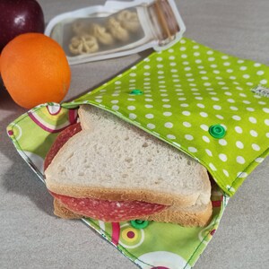 Könnte beinhalten: Eine wiederverwendbare Sandwich-Tasche mit grünen und weißen Punkten, in der sich ein Salami-Sandwich befindet. Die Tasche hat einen grünen Knopfverschluss.