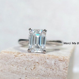May include: A silver ring with an emerald cut diamond. The ring is on a white stone surface. Merci Ma Belle Store is written in the background.