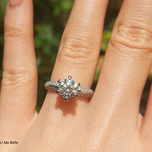 May include: A silver engagement ring with a large, round, clear diamond center stone and smaller diamonds set in a pave setting around the center stone. The ring is on a person's finger.