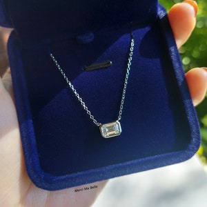 May include: A silver chain necklace with a small, clear, emerald-cut gemstone pendant. The necklace is in a blue velvet box with the words "Merci Ma Belle" printed on the bottom.