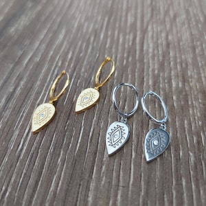 May include: Three pairs of earrings with a teardrop-shaped charm featuring an evil eye design. The charms are gold and silver with a clear rhinestone in the center of each eye.