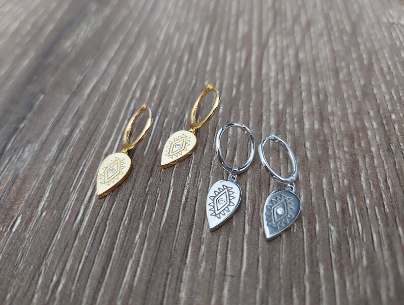 May include: Three pairs of earrings with a teardrop-shaped charm featuring an evil eye design. The charms are gold and silver with a clear rhinestone in the center of each eye.