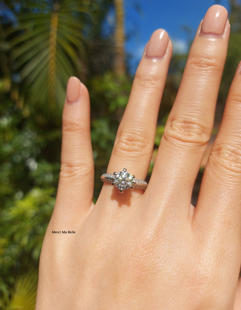 May include: A silver engagement ring with a large, round, clear diamond center stone and smaller diamonds set in a pave setting around the center stone. The ring is on a person's finger.