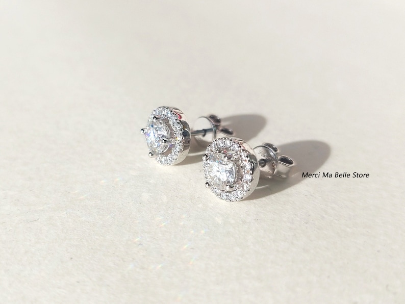 May include: A pair of silver stud earrings with a round, clear crystal center surrounded by a halo of smaller crystals.