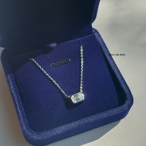 May include: A silver necklace with a rectangular, clear gemstone pendant in a blue velvet box. The box has the text "Merci Ma Belle" printed on it.